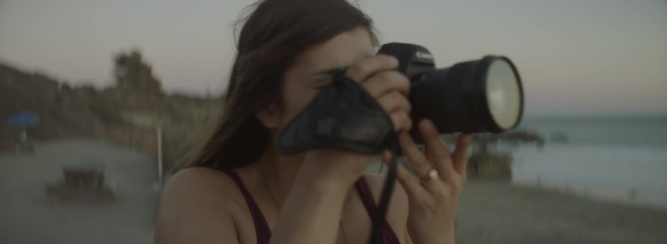 A woman raises a camera to her eyes while standing on a beach