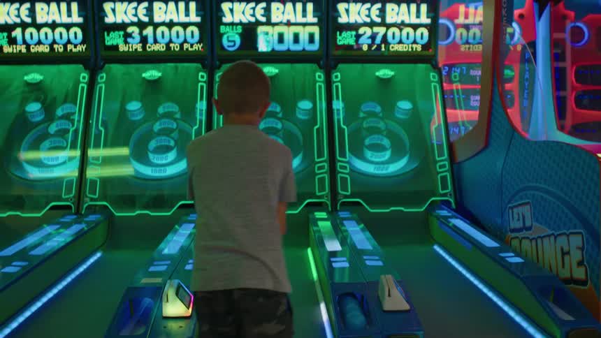 A boy plays skee ball in an arcade with lights flashing and glowing