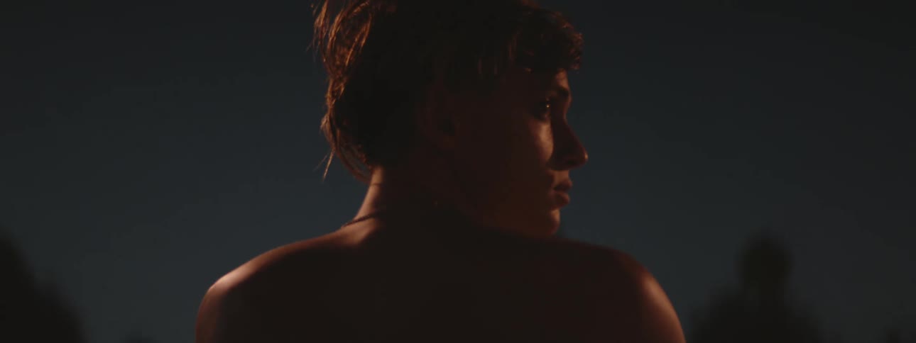 A shirtless man outside at night turns his head side to side in a nervous manner