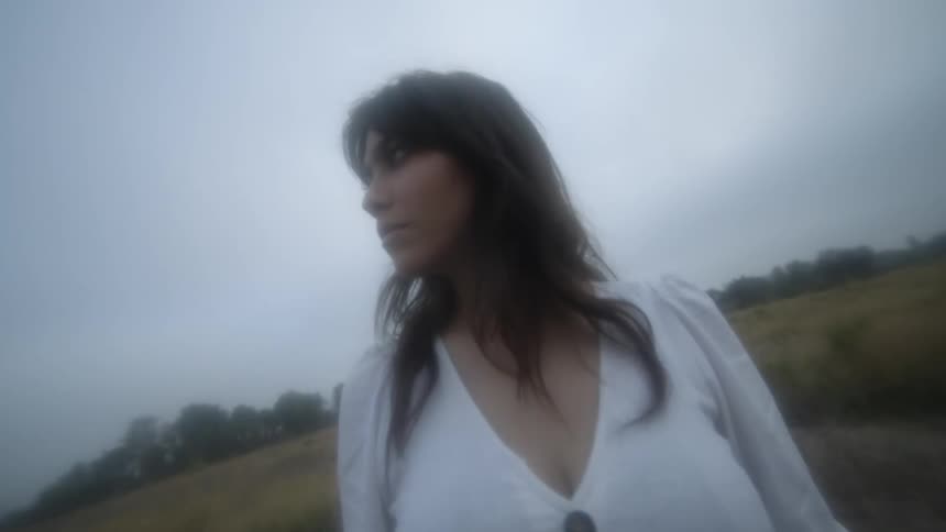 Woman stands outdoors looking around under overcast sky near field of brownish yellow grass