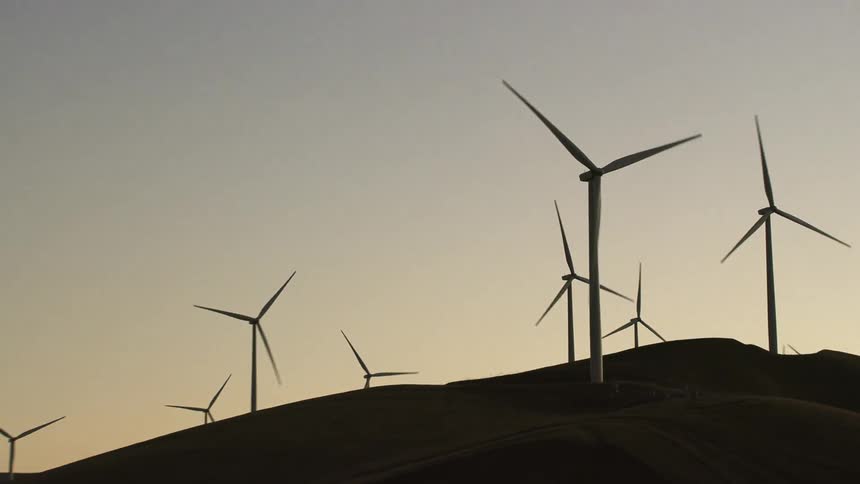 Turbine blades rotate in wind farm set on hill against red tinted sky