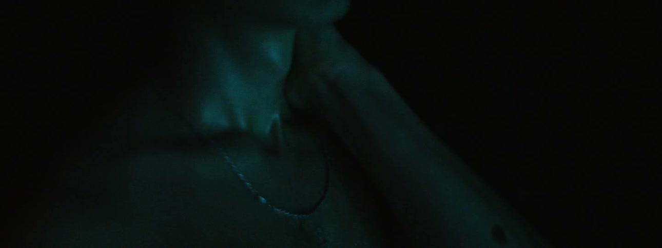 A shirtless man touches his chain and medallion as his skin glistens in a passing light
