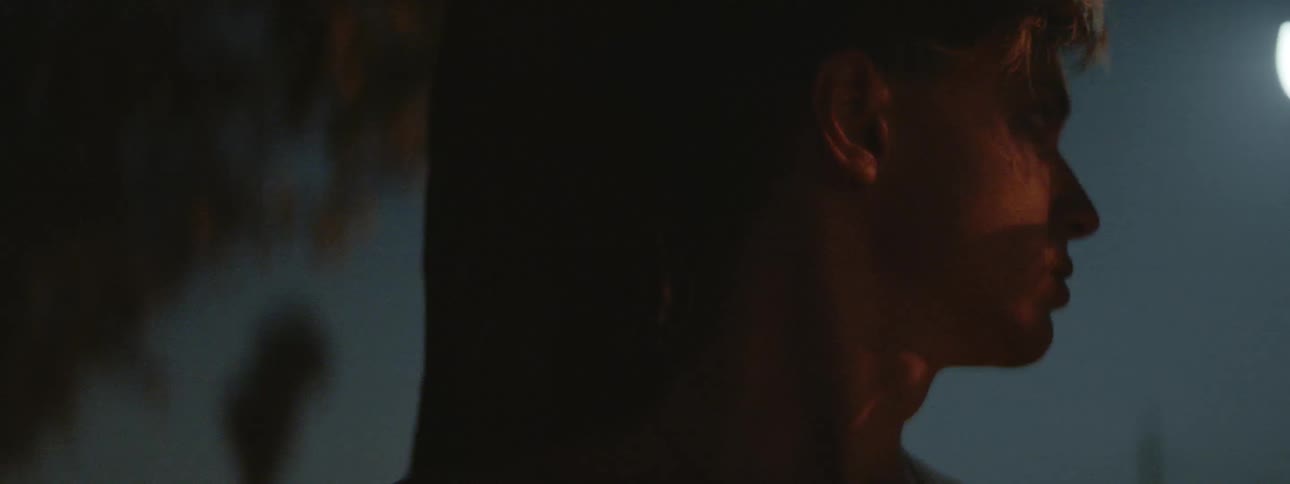 A teenage boy stands against a tree at night and worriedly looks around his surroundings