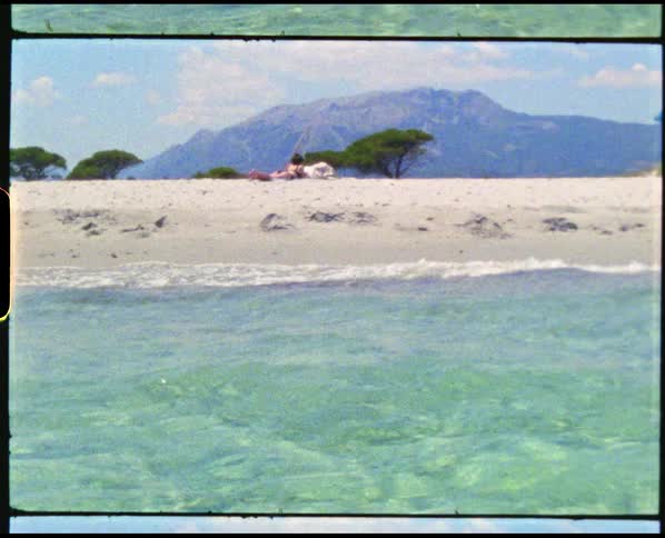 Woman sunbathes on beach near clear blue water with snow covered mountain in background under clouded sky