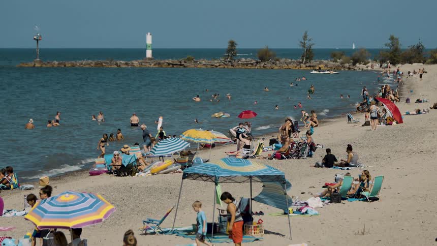 A large amount of people sit and walk through the beach sand as well as stand in the ocean water