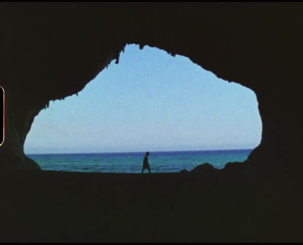 Person walking across mouth of sea cave is silhouetted against blue of ocean waves