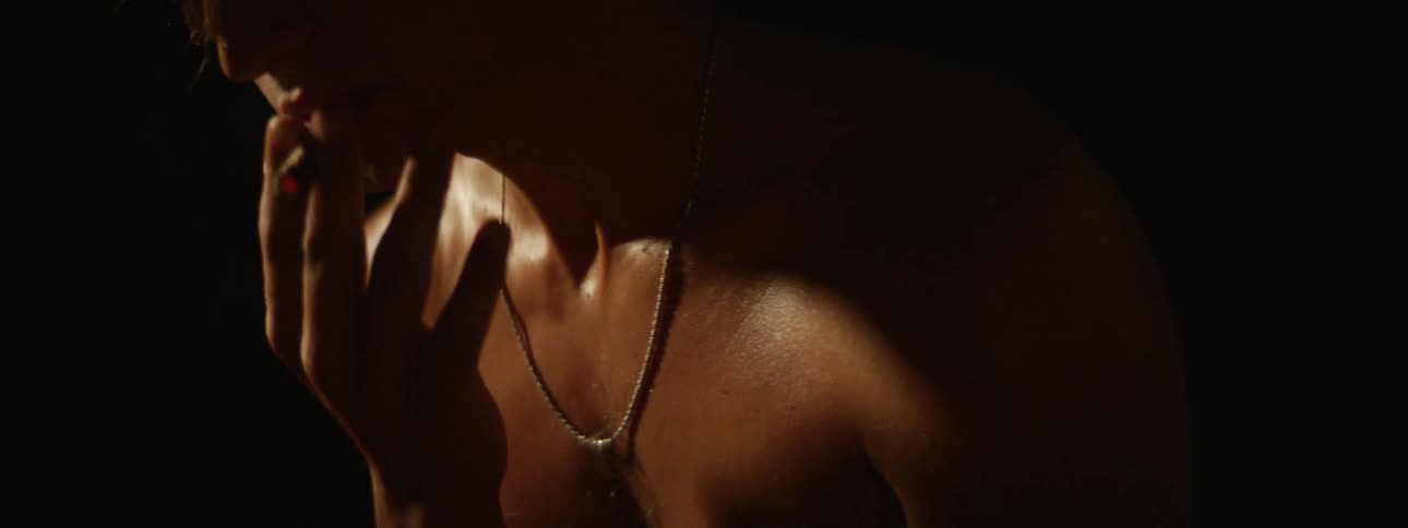 A shirtless man wearing a chain smokes with light shining on his glistening skin