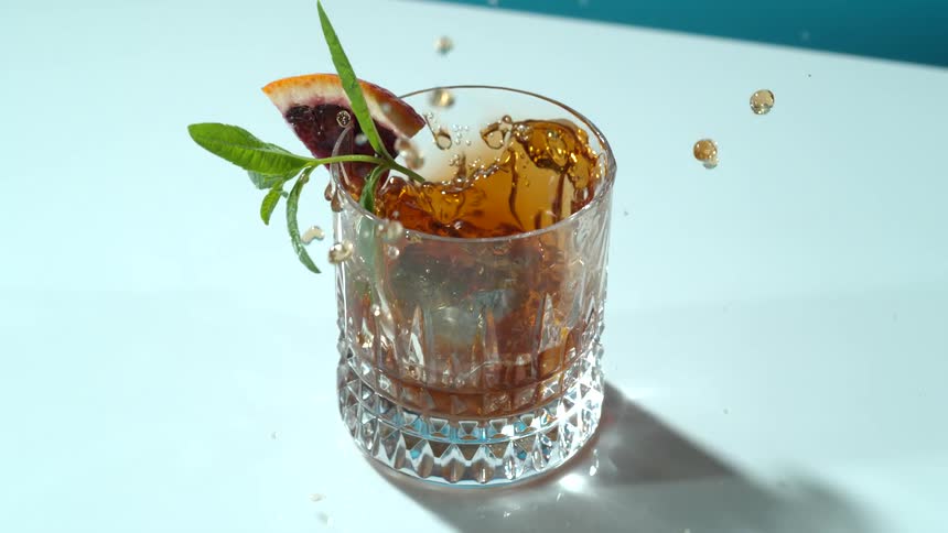 A drink in a room on a table with a cement cube falling into the drink with a wall in the background