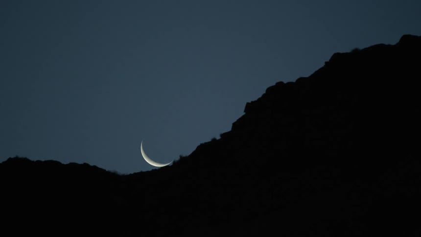 Crescent moon sets behind hill saddle in clear sky at night