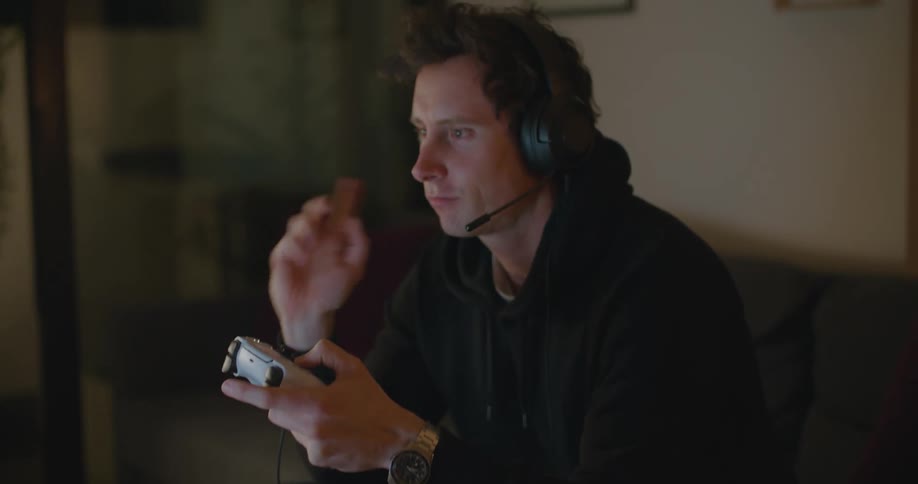 A man sits on a couch playing a video game and gesturing while talking into a headset