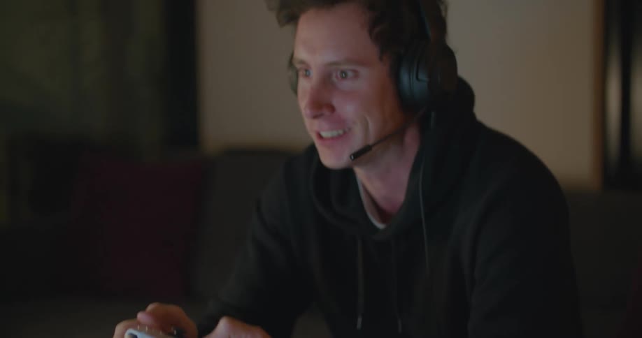 Man wearing hoodie sits on couch manipulating game controller smiles and yells