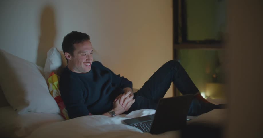 A man in a room laying on a bed looking at a laptop with a wall and window in the background