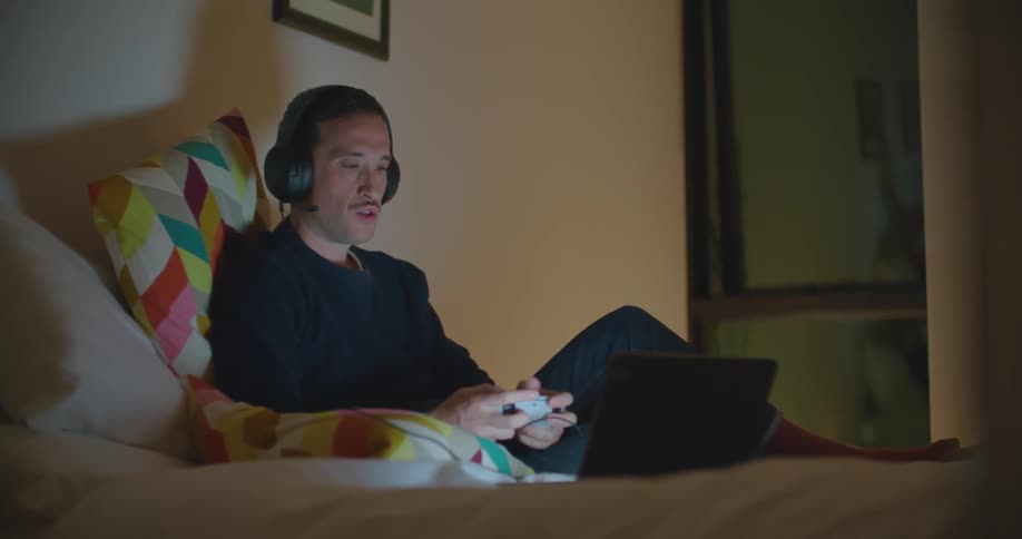 Man sits on bed in dimly lit room playing game using controller attached to laptop
