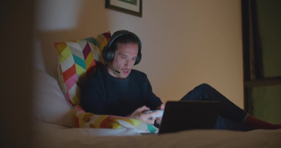 Man sits on bed in dim bedroom playing video on laptop using game controller and wearing gaming headset