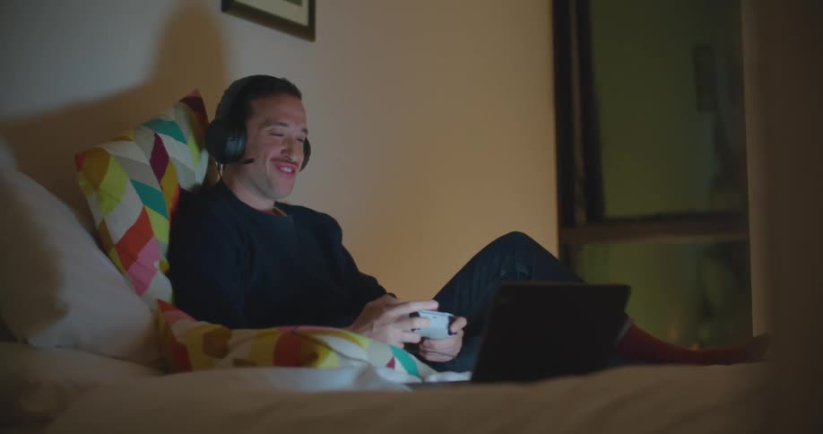 A man with a headset and a videogame control looking at a laptop in a room with a wall and window in the background