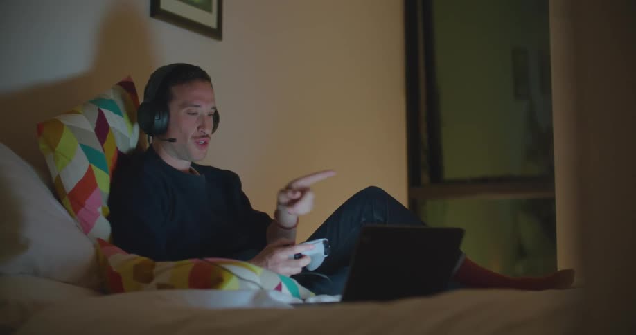 A man sits on a bed playing a game on a laptop holding a controller and talking into a headset