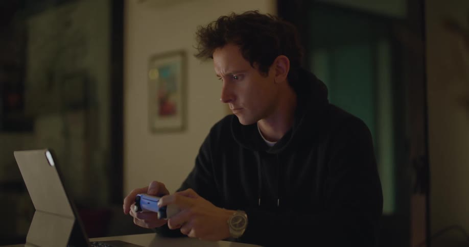 A man uses a gamepad to play a video game on a computer tablet while sitting at a table indoors
