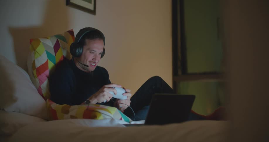 A man sits on a bed and plays a video game on a laptop while talking on a headset