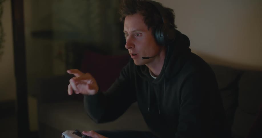 A man is sitting in a room playing a video game and talking to someone on a headset