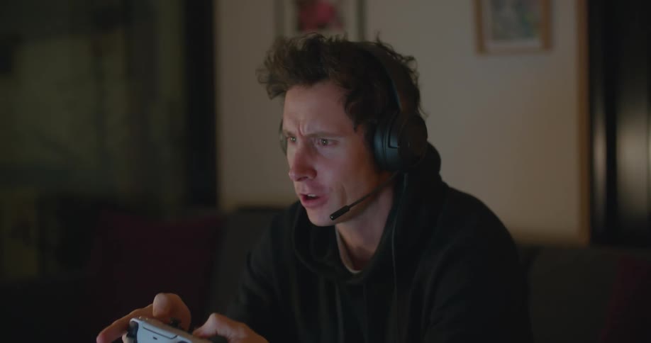 A young man sits at home and talks in to a headset while playing a video game