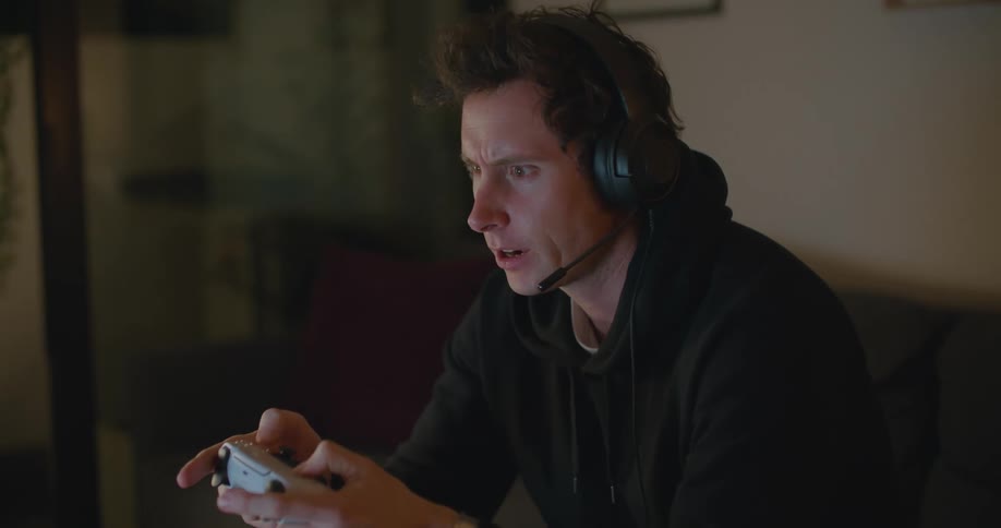 A man grimaces as he plays a video game and talks on a headset