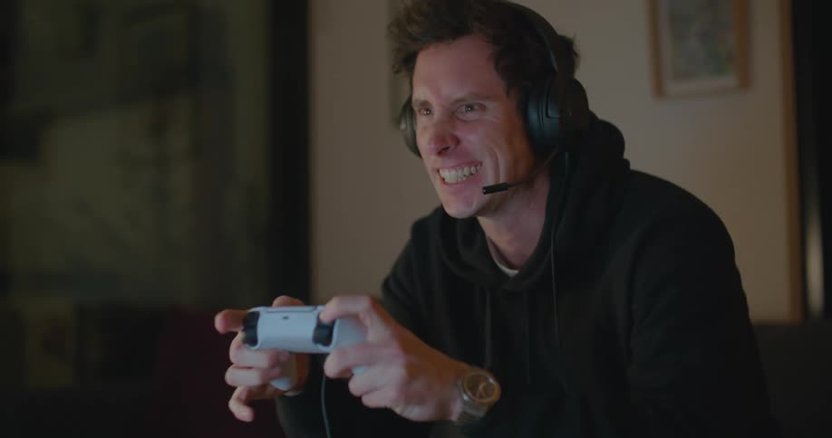 A man sits and plays a game with a controller in his hand and talking in a microphone with headphone