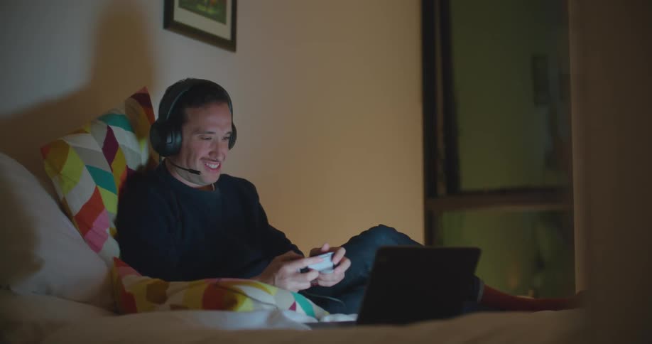 A man sits on a bed playing a video game on a laptop and talking into microphone headphones