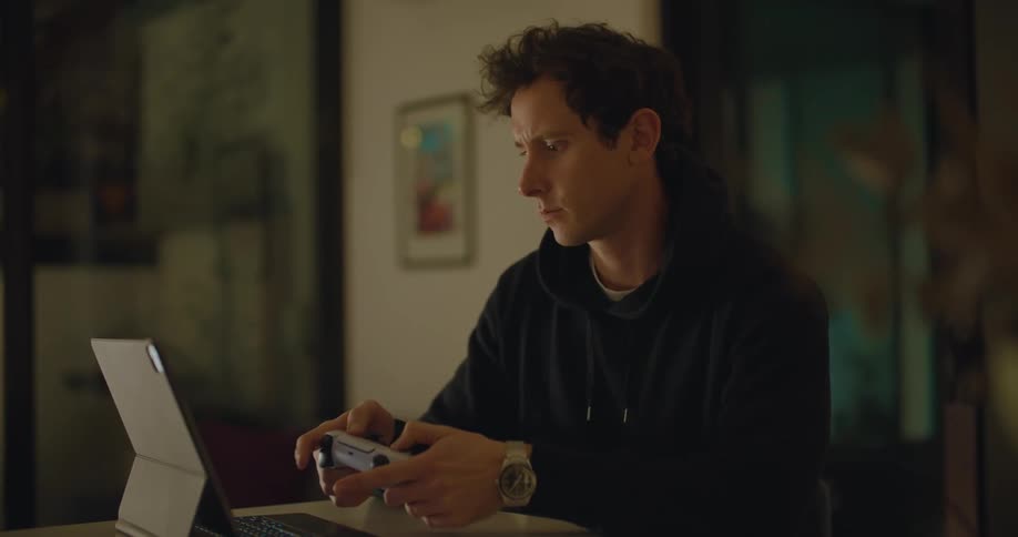 A man in a room with a videogame controller looking at a tablet with a wall and door in the background