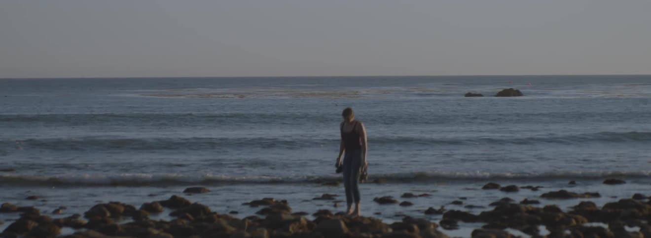 A woman holding her shoes and a camera stands amidst tide pools on a beach