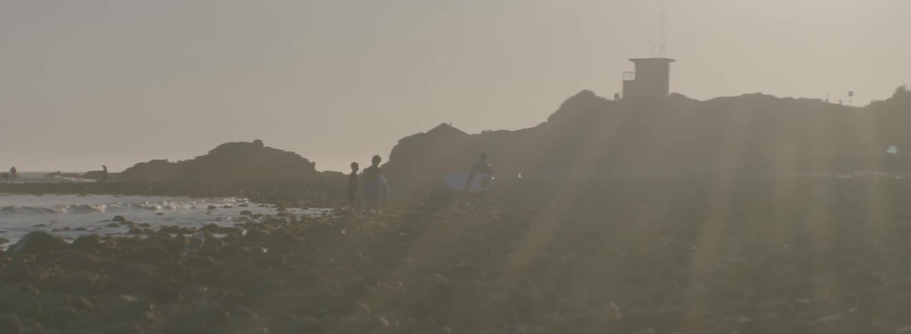 People are walking on the rocks walking towards the water with a surfboard