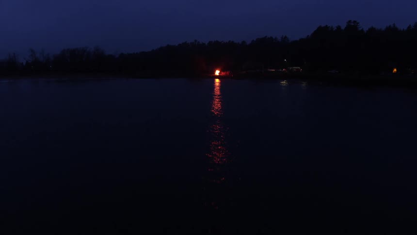 Bonfire burning on shore reflects off rippling water brightening area surrounded by woods