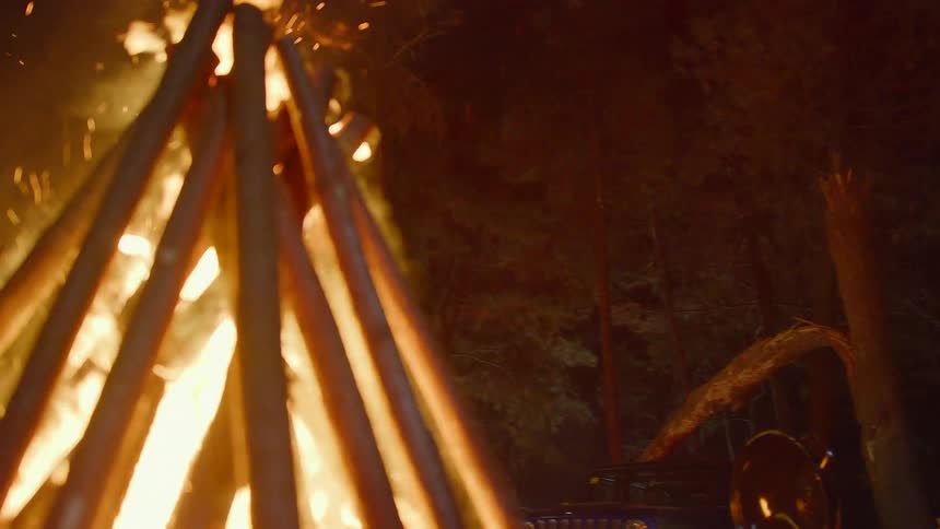 Embers fly from a flaming campfire with a tent near the trees