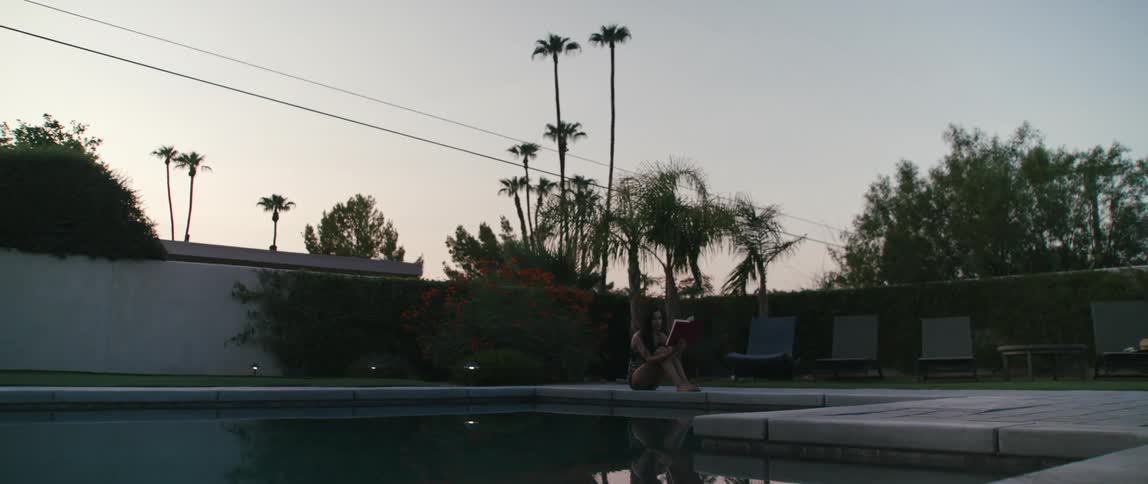 A woman sits on a pool deck reading a book with sky and trees reflected in the water surface