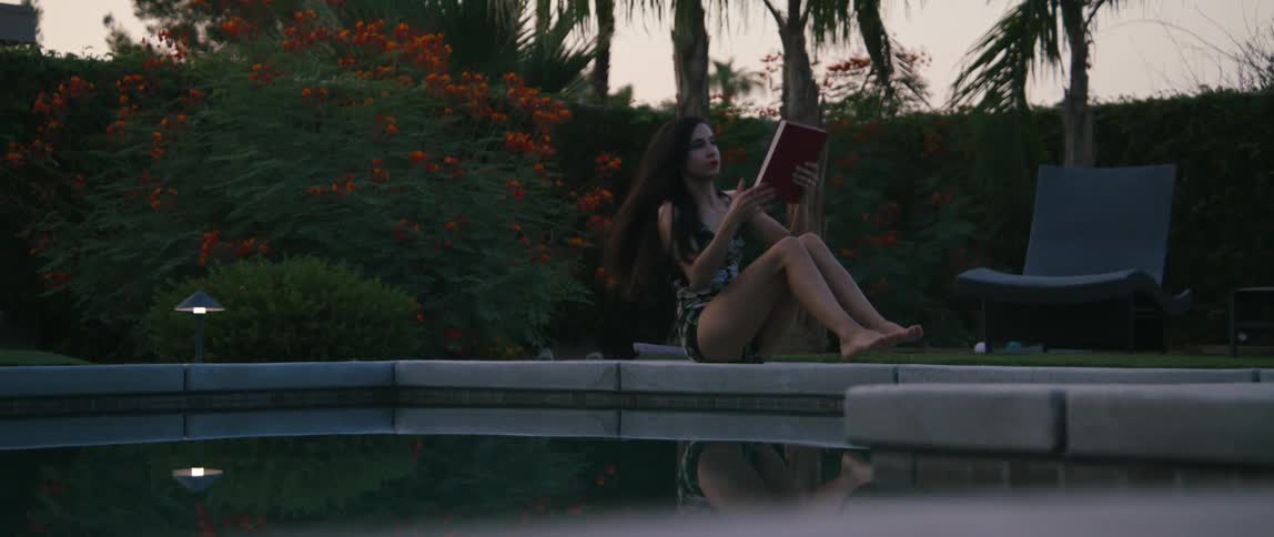 A woman sits down beside a pool and opens a book to read