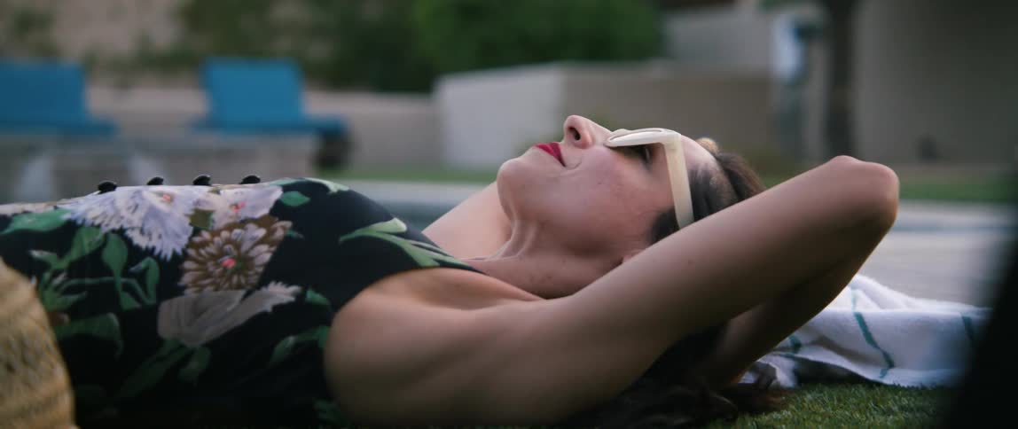 A woman with sunglasses laying down on grass with a wall and trees in the background