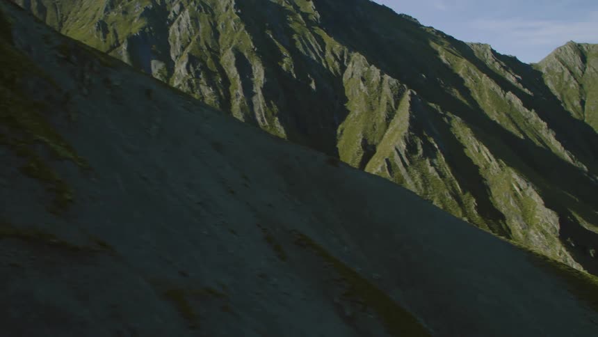 The camera passes by a rocky range of mountains that are covered in green vegetation