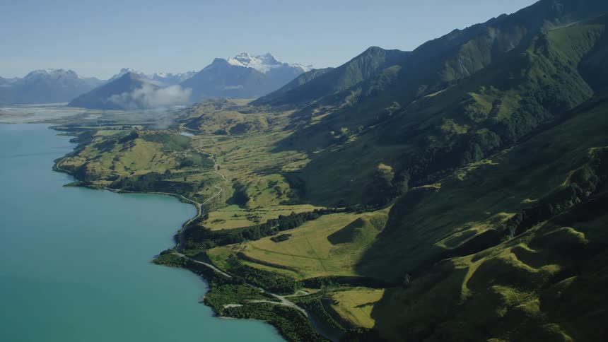 A verdant mountain slope leading down to a road beside the coast during the day