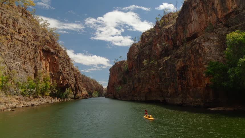 Two people in a yellow boat row down a river in a canyon on bright blue sky day