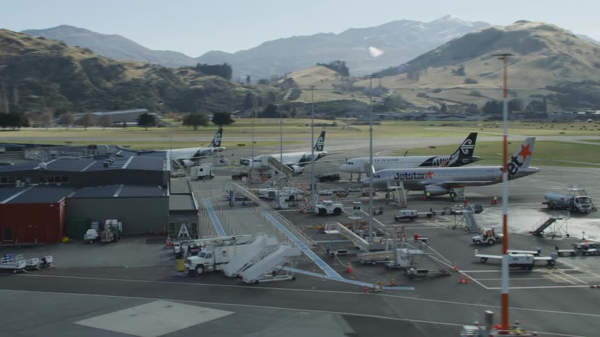 Airport employees move around airliners sitting on tarmac with buildings and hills in distance