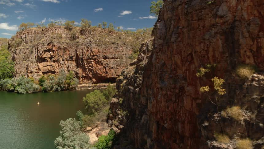 A person rows a yellow kayak on a river flowing between rocky cliffs on a bright sunny day