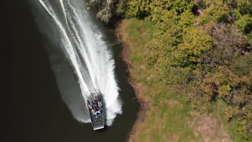 An aerial view of people in a speedboat driving along the shore of a river