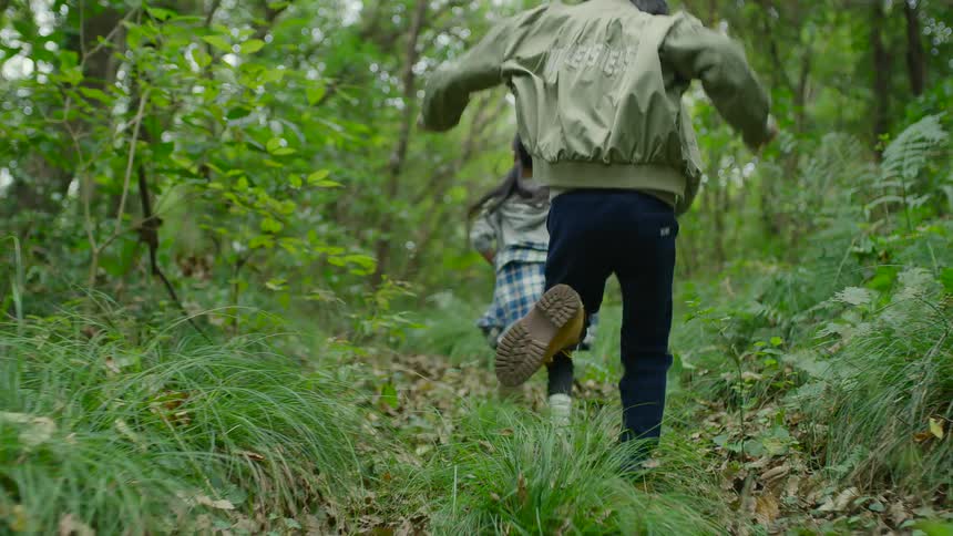A girl and boy run together through a forest during the day