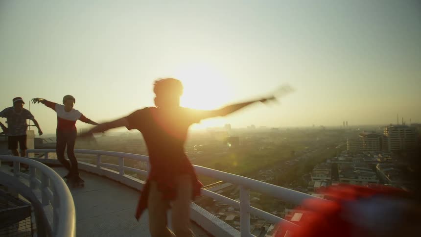 A group of boys raise their arms in excitement as they skateboard across a narrow high bridge with the city below