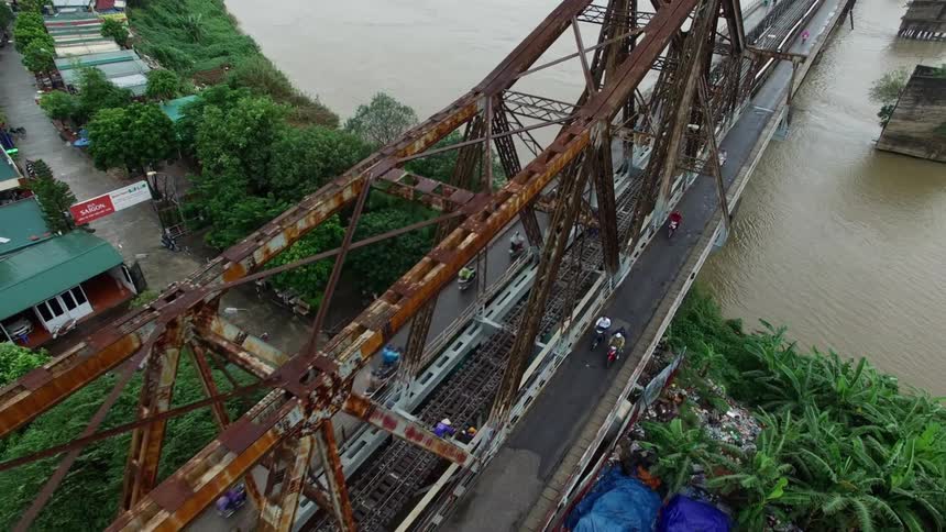 Motorcyclists and pedestrians cross rusted bridge over muddy water bordered by houses on one side and canvas roofed shacks on the other