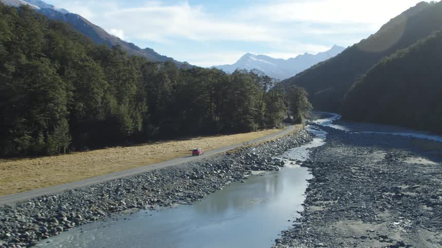 A vehicle travels down a countryside road between a forest and a stream