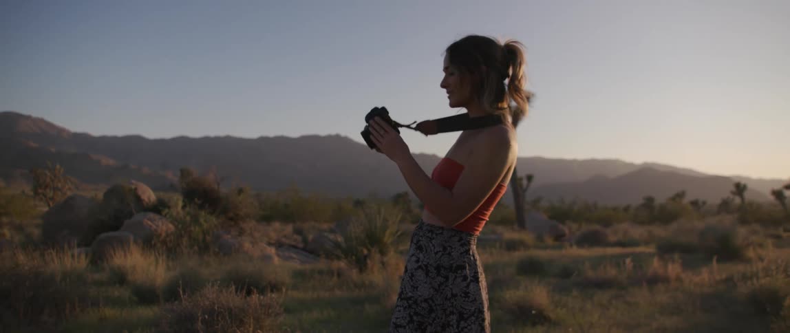 Woman with red sleeveless shirt and patterned skirt takes photographs on shrubland at sunset
