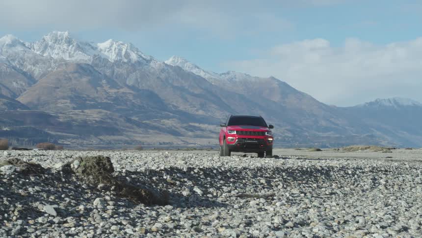 A red sport utility vehicle drives over rockcovered terrain near to a mountain range