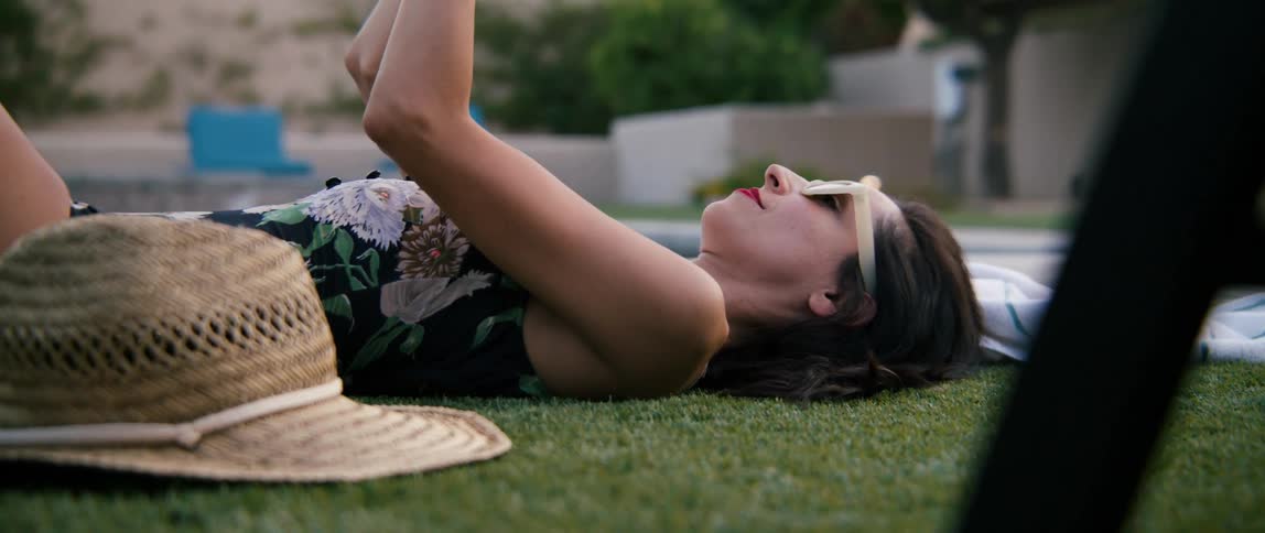 Woman lies on back on turf near pool reading book