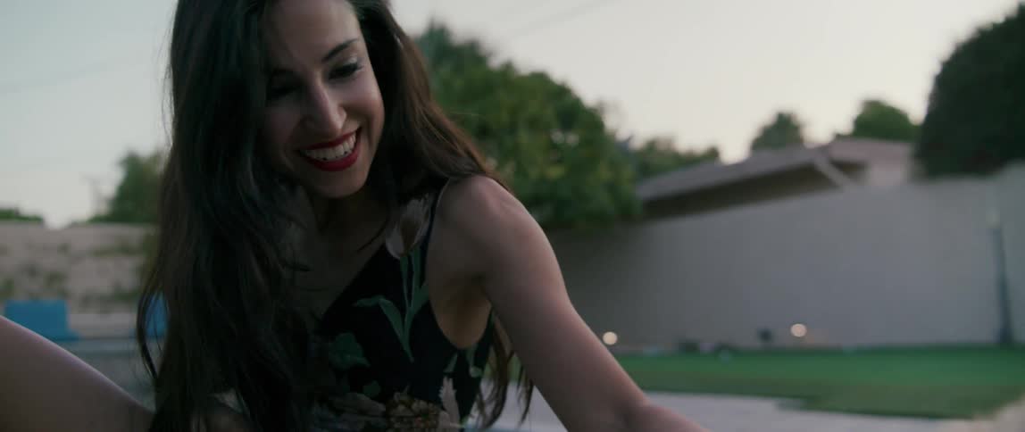 A woman by a pool petting a dog with a wall trees the sky and clouds in the background