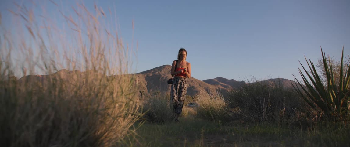 A woman carrying a camera through a grassland in front of a mountain range